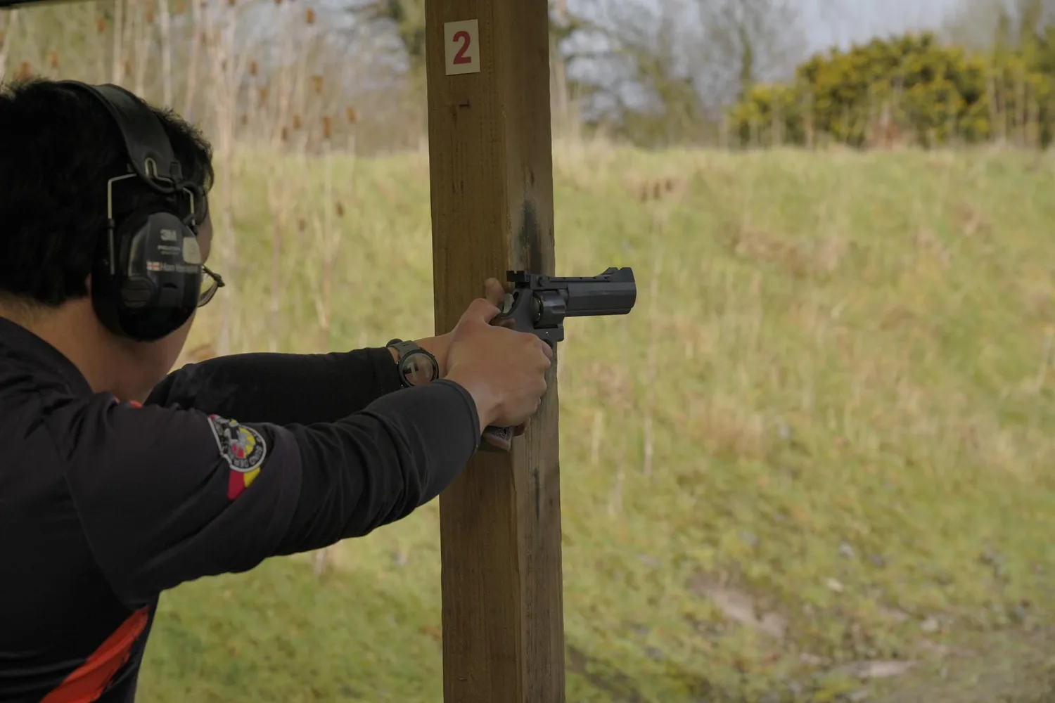 A pistol shooter engaging a target from behind a barricade on a range, wearing a Garmin watch on his wrist.
